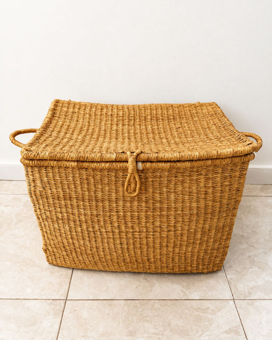 A rectangular woven storage basket with a lid, made from natural tan seagrass or wicker, featuring integrated side handles and a rope loop closure on the lid, set against a plain white wall and light beige tiled floor.