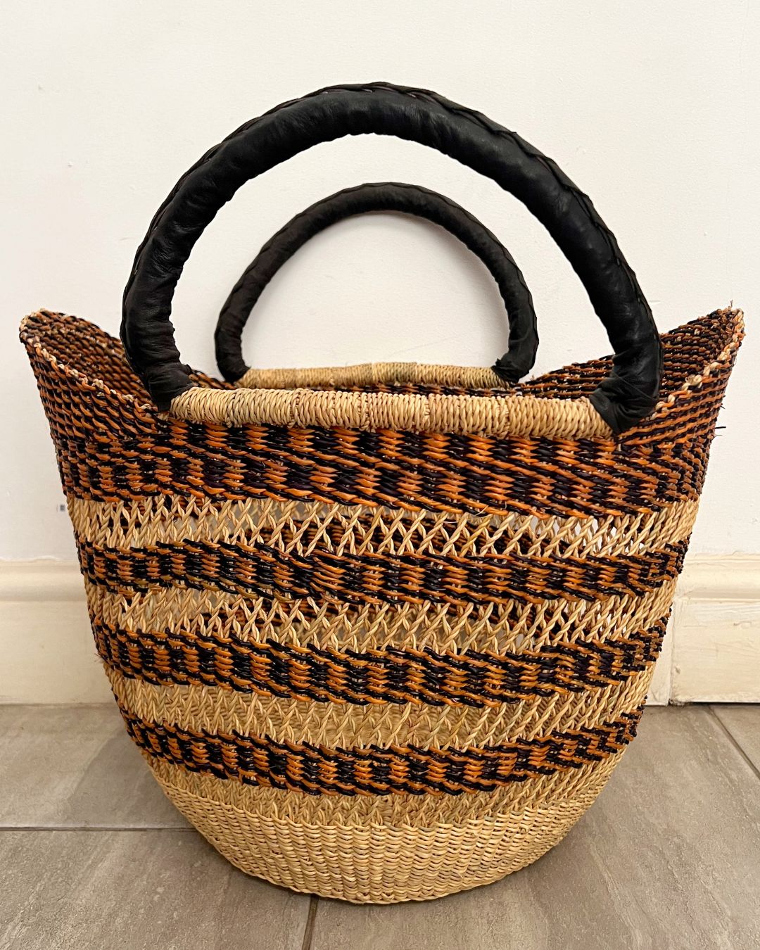 A woven straw tote bag with black handles, featuring horizontal stripes of natural beige, brown, and black raffia on a light gray floor against a white wall.