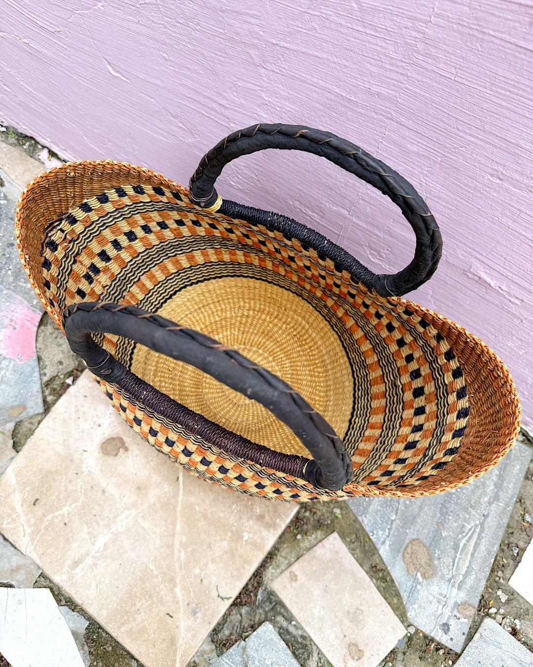 A woven straw basket with black and brown braided handles, photographed from above on a stone surface against a light purple wall.