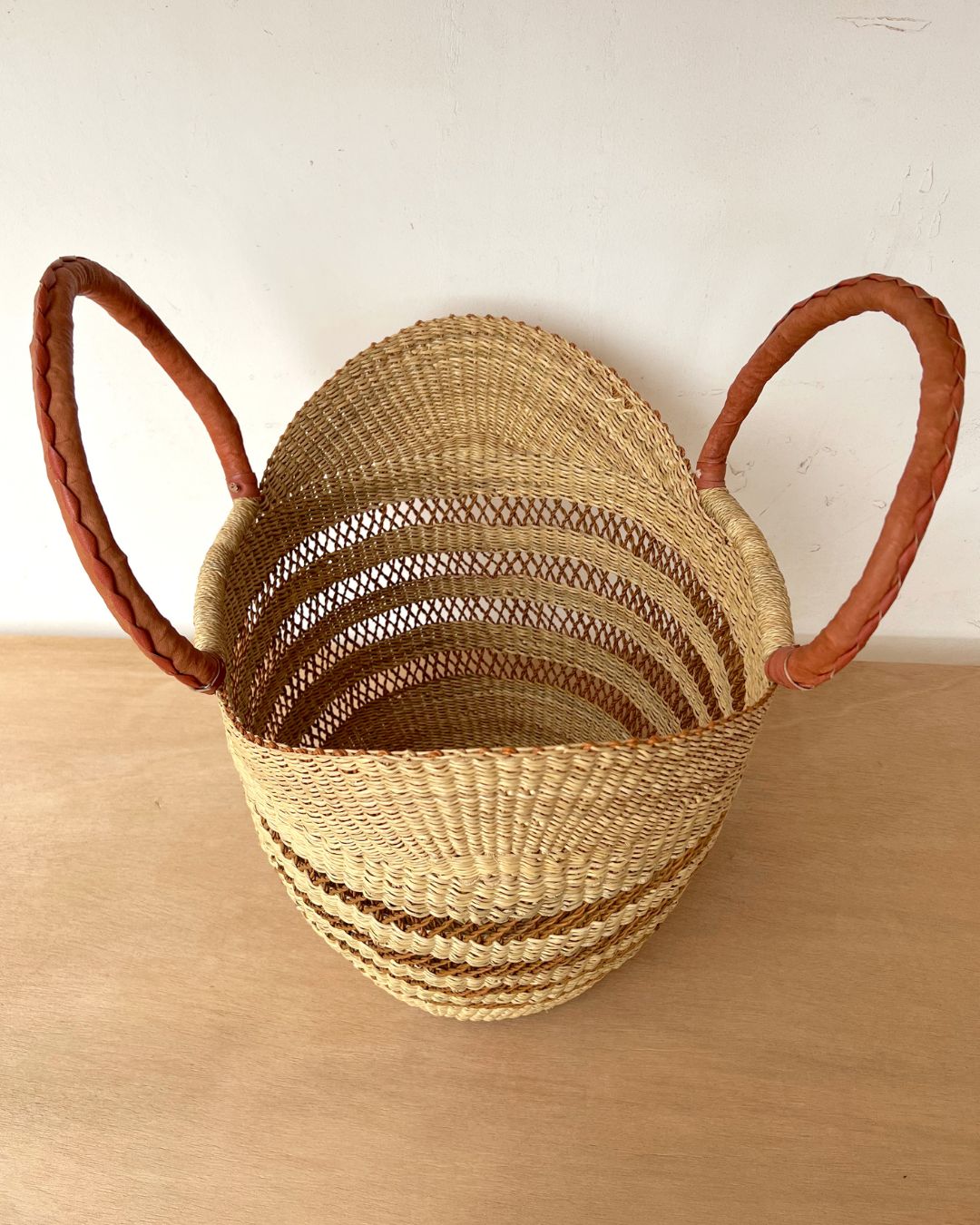 A natural woven straw basket with two braided brown handles, shown on a light wood surface against a plain off-white wall.
