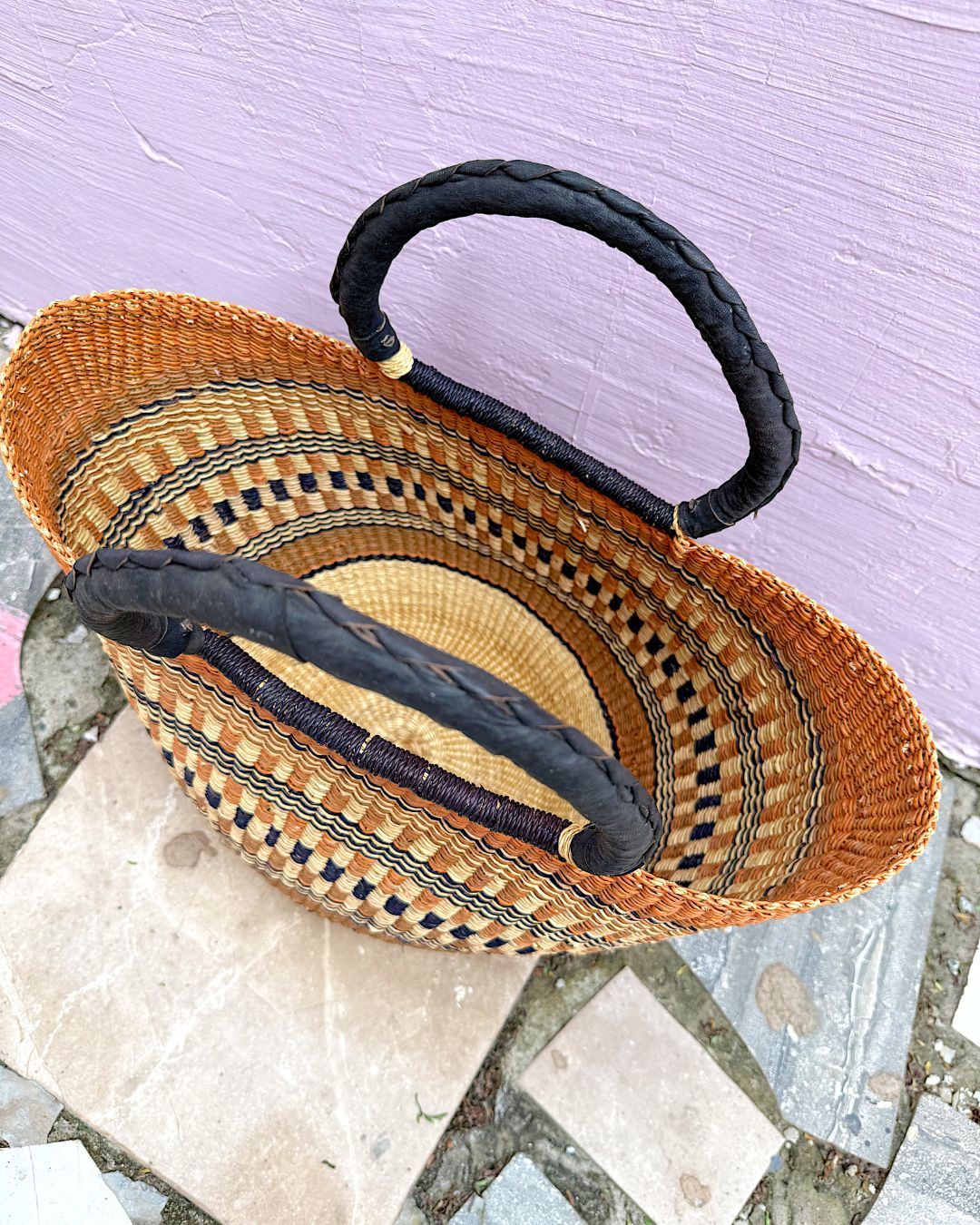 A woven straw tote bag with black braided handles, photographed from above on stone tiles against a light purple wall.
