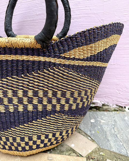 A close-up of a woven basket bag with dark navy and natural tan stripes, featuring dark brown leather handles, set against a light pink wall and stone floor.