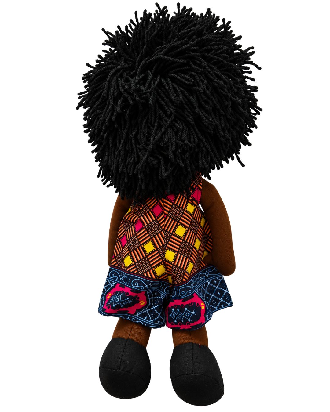 A plush doll viewed from the back, featuring a large black textured afro hairstyle, brown skin, a yellow, red, and black patterned top, and blue patterned shorts with pink accents, set against a plain white background.