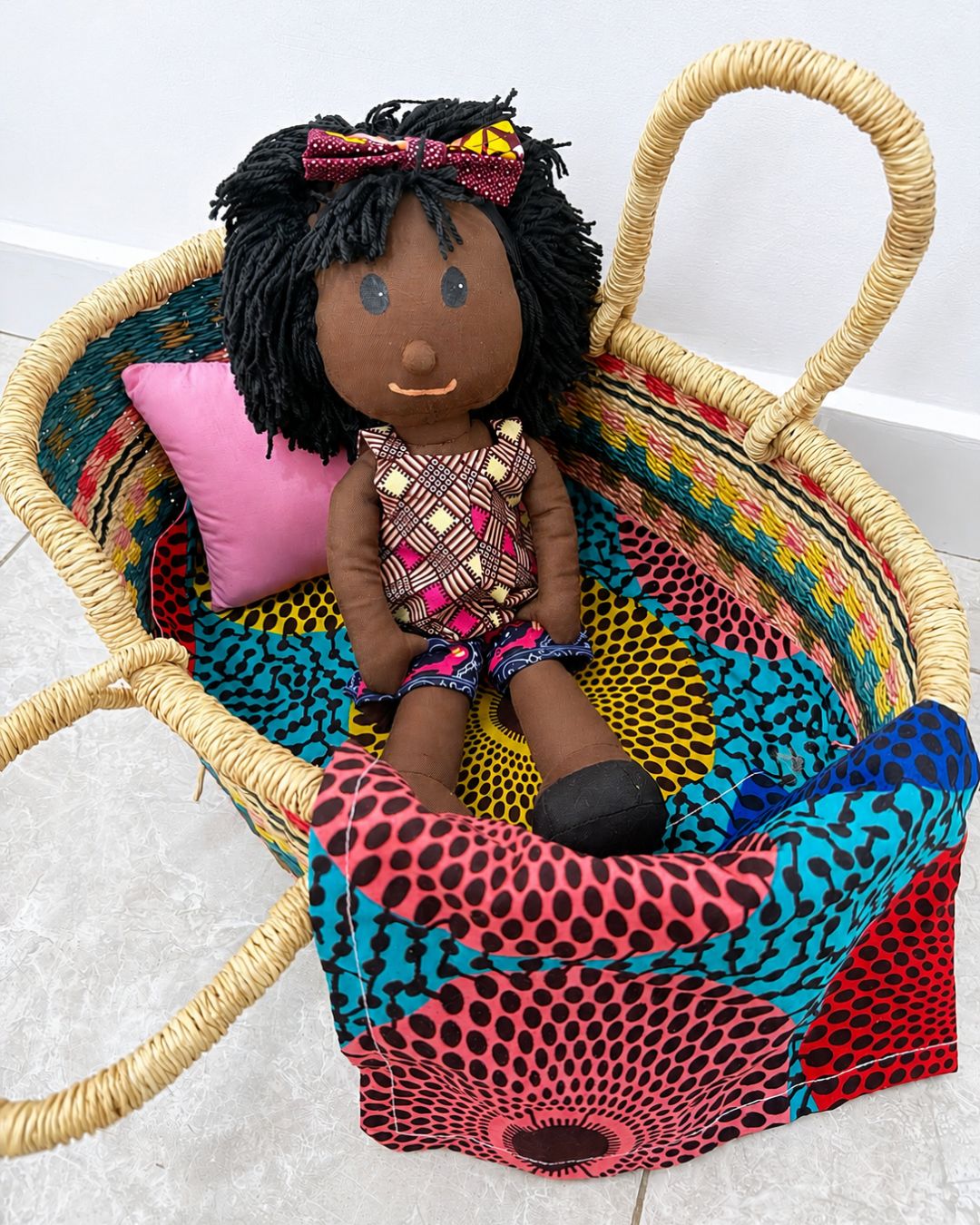 A handmade African-style woven basket with a colorful patterned fabric lining and a pink pillow, containing a handmade African doll with black yarn hair and a pink bow.