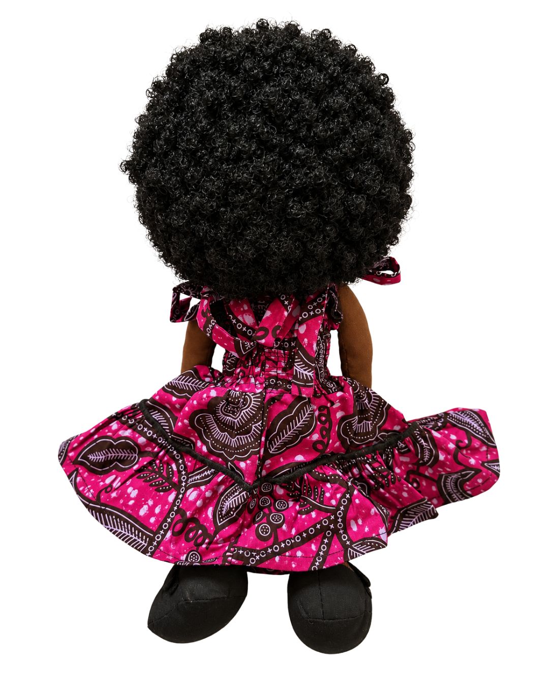 A doll viewed from the back, featuring a large black afro wig, dark brown skin, and a pink dress with a black and white paisley pattern, set against a plain white background.