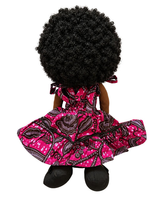 A doll viewed from the back, featuring a large black afro wig, dark brown skin, and a pink dress with a black and white paisley pattern, set against a plain white background.