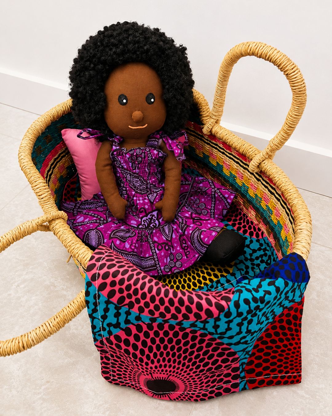 A handmade woven basket with a colorful patterned fabric liner and a pink pillow, containing a plush doll with black hair and a purple patterned dress, set against a plain white wall and light floor.