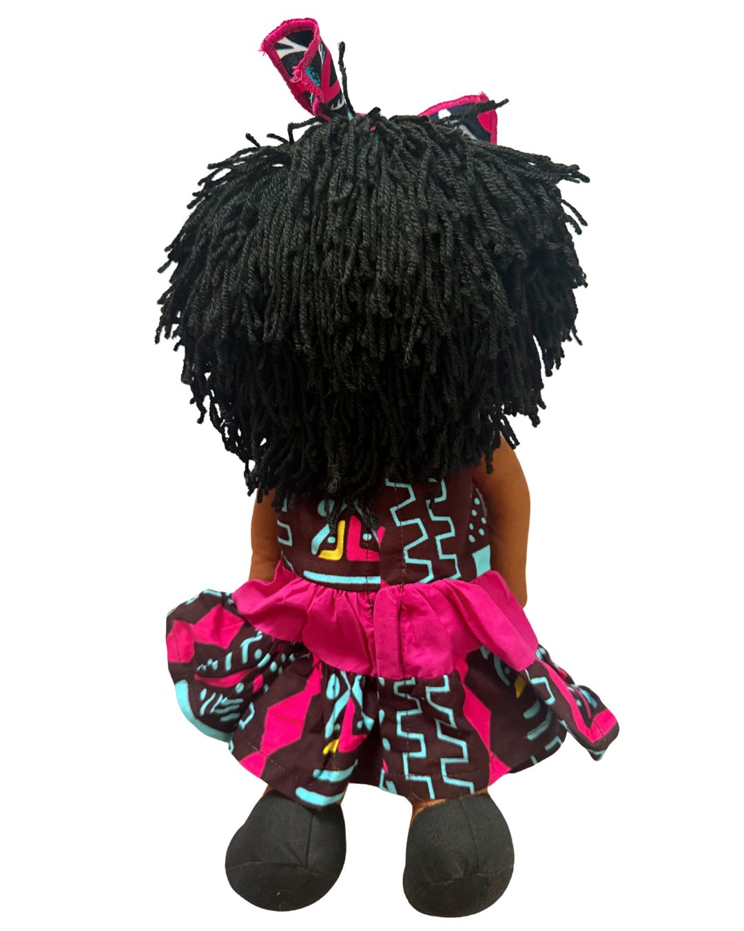 A doll viewed from the back, featuring a large black afro wig with a matching pink and black hair bow, a patterned pink, black, and turquoise dress, and black shoes, isolated on a white background.
