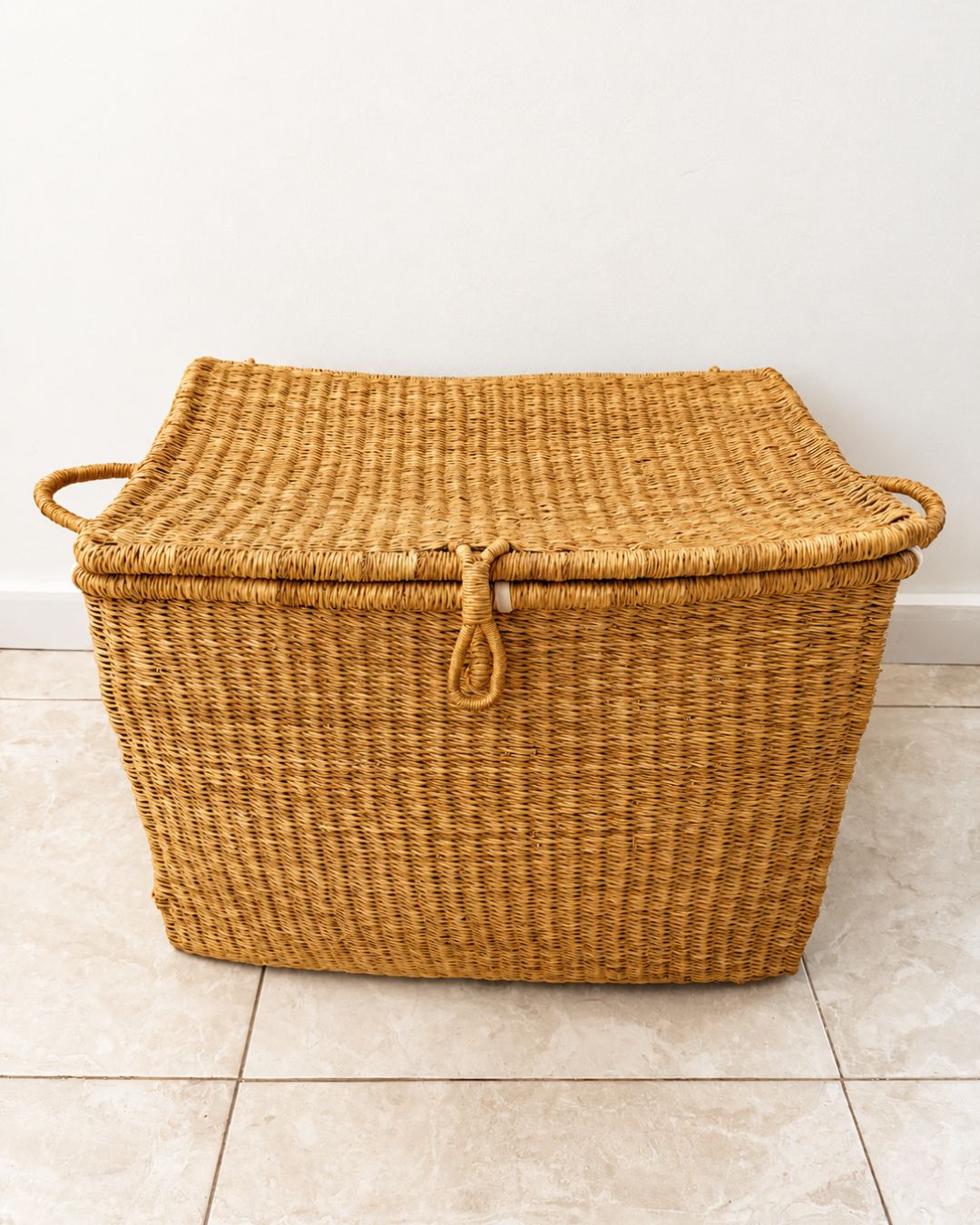 A rectangular woven storage basket with a lid, made from natural tan seagrass or wicker, featuring integrated side handles and a rope loop closure on the lid, set against a plain white wall and light beige tiled floor.