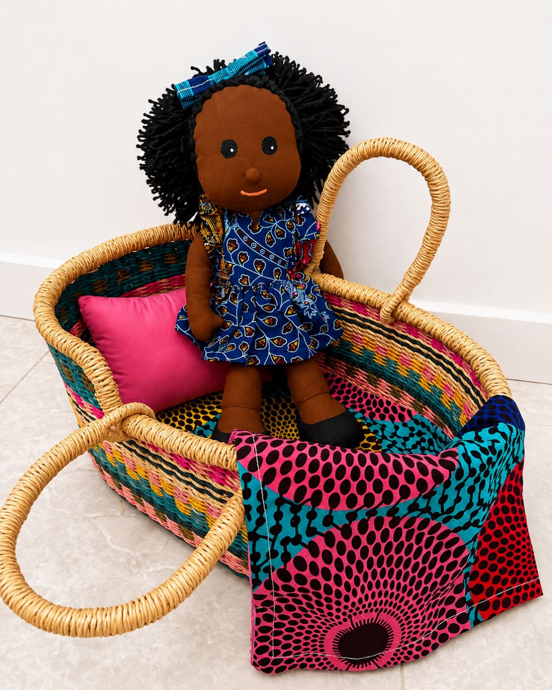 A colorful woven basket with a doll sitting inside, styled with a pink pillow and patterned fabric on a light tiled floor against a white wall.