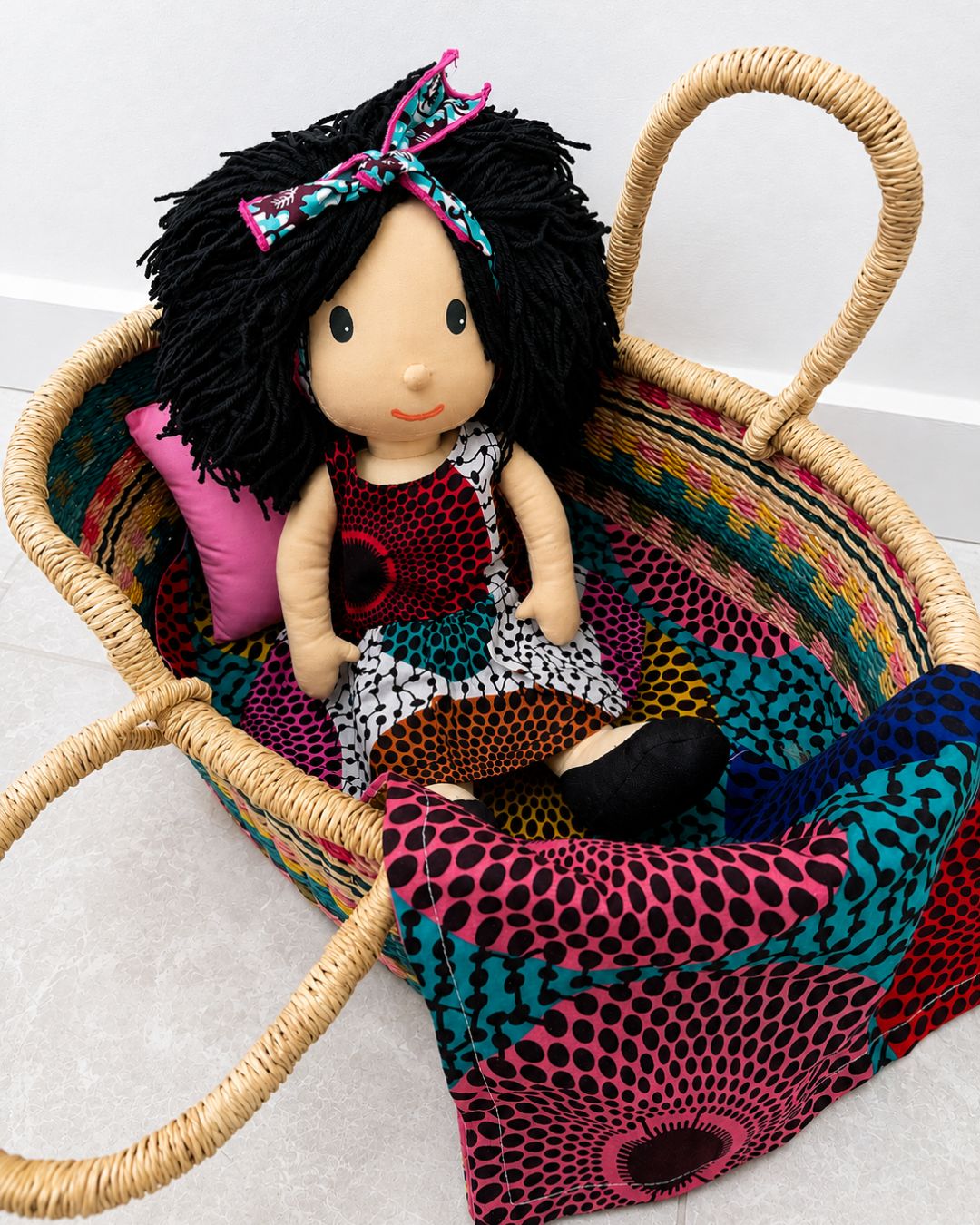 A handmade fabric doll with black yarn hair and a colorful patterned dress sits inside a woven straw basket lined with matching multicolor patterned fabric.