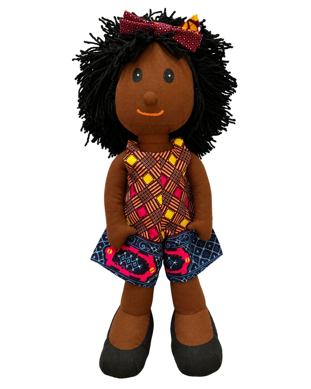 A handmade African American doll with black yarn hair, a red polka-dot bow, and a colorful patterned dress and shorts, shown against a plain white background.
