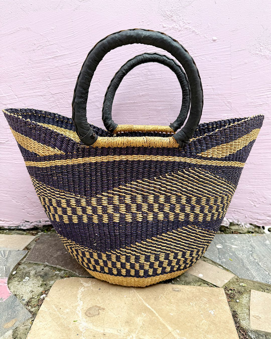 A woven basket-style tote bag with dark navy and tan geometric stripes, featuring two thick black loop handles, set against a light pink wall and stone floor.