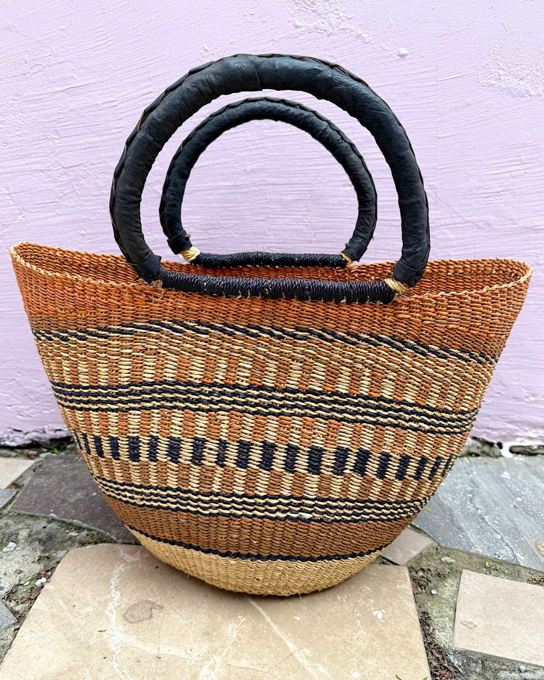 A woven straw tote bag with black circular handles, featuring horizontal stripes of black, brown, and beige, set against a light purple wall.