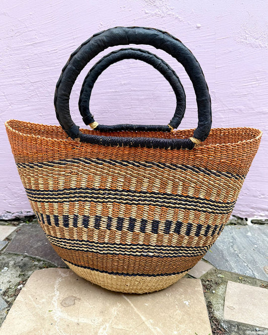 A woven straw tote bag with black circular handles, featuring horizontal stripes of black, brown, and beige, set against a light purple wall.