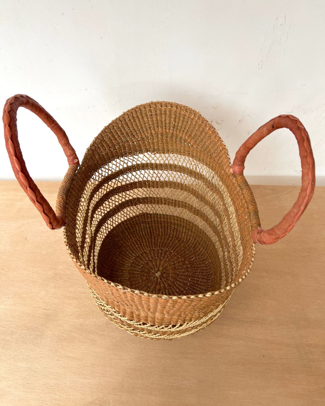 A natural woven basket with two curved reddish-brown handles, shown on a light wood surface against a plain off-white wall.