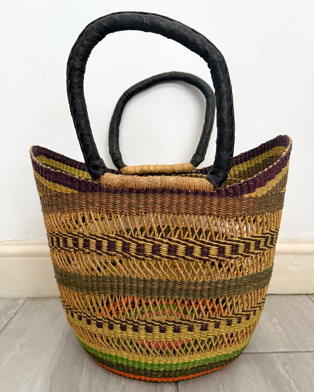 A hand-woven basket bag with a multicolor striped pattern in natural tan, olive green, yellow, and purple, featuring two thick black fabric handles, set against a plain white wall and light gray floor.