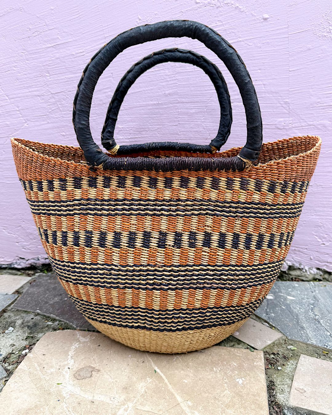 A woven straw tote bag with black and tan horizontal stripes, featuring two thick black loop handles, set against a light purple wall and stone tiles.