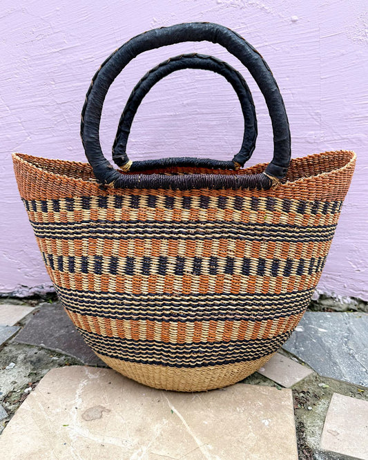 A woven straw tote bag with black and tan horizontal stripes, featuring two thick black loop handles, set against a light purple wall and stone tiles.