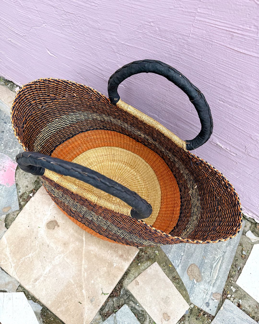 A hand-woven basket with dark brown and black woven body, black handles, and orange and beige striped inner lining, resting on stone tiles against a lavender wall.