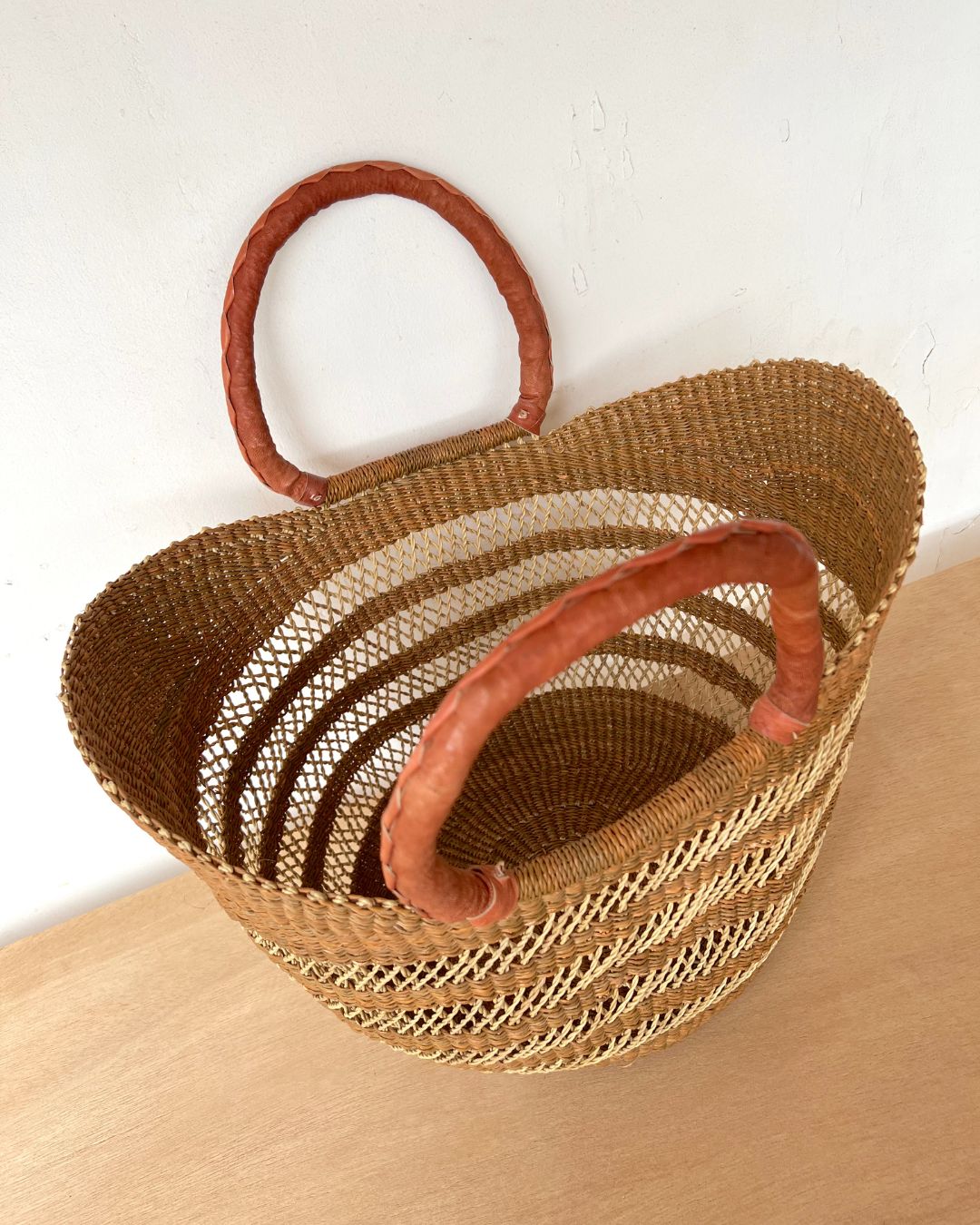 A woven straw tote bag with a rounded shape, featuring two thick, twisted brown handles and a natural beige and light brown striped pattern, resting on a light wood surface against a white wall.