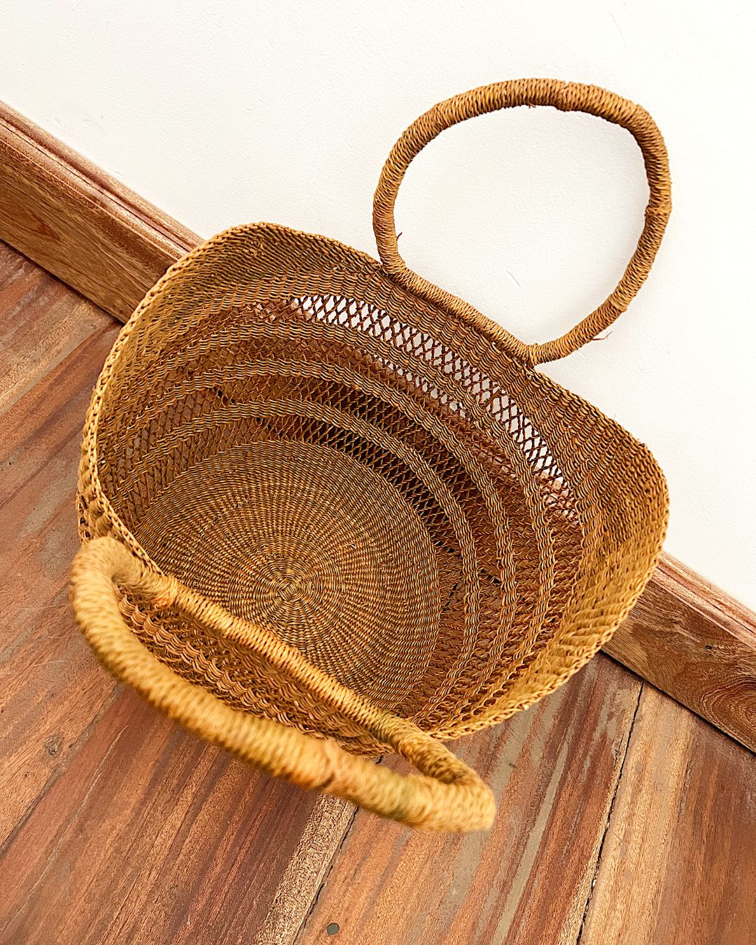 A natural woven straw basket with two loop handles, photographed from above on a wooden floor against a white wall.
