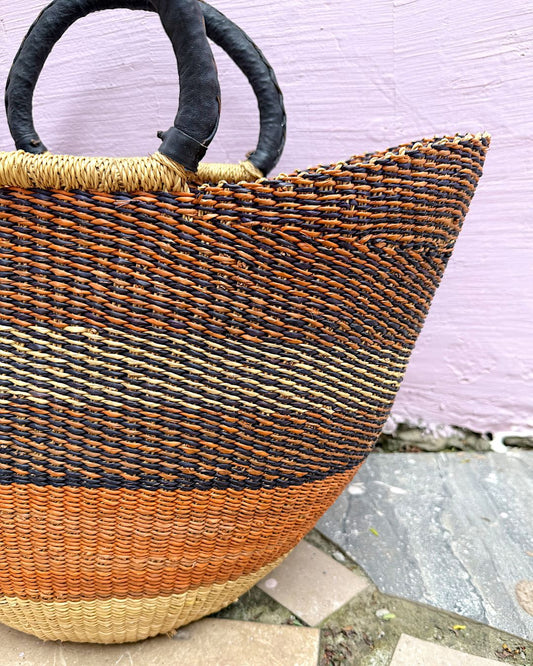 A woven straw tote bag with black handles, featuring horizontal stripes of black, tan, and orange-brown, set against a light purple wall.
