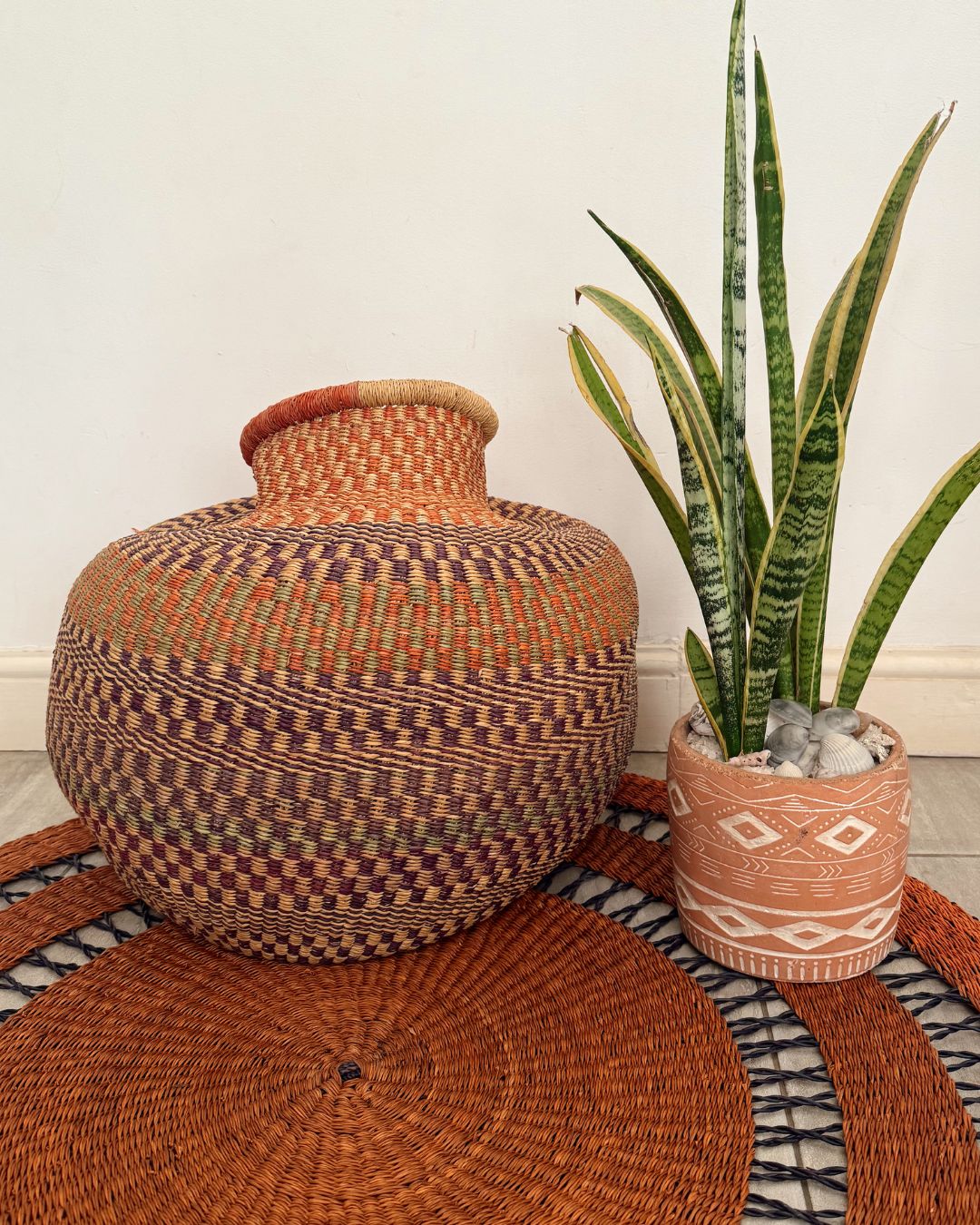 A large round hand-woven basket vase in earthy brown, orange, and purple tones, styled next to a potted snake plant on a woven rug against a plain wall.