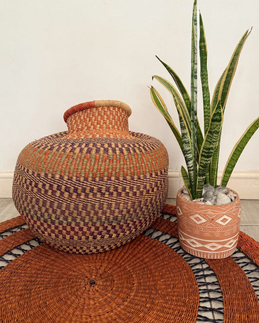 A large round hand-woven basket vase in earthy brown, orange, and purple tones, styled next to a potted snake plant on a woven rug against a plain wall.