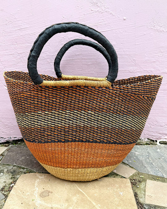A hand-woven straw tote bag with a striped pattern in brown, black, and orange, featuring two thick black woven handles, set against a light pink textured wall.
