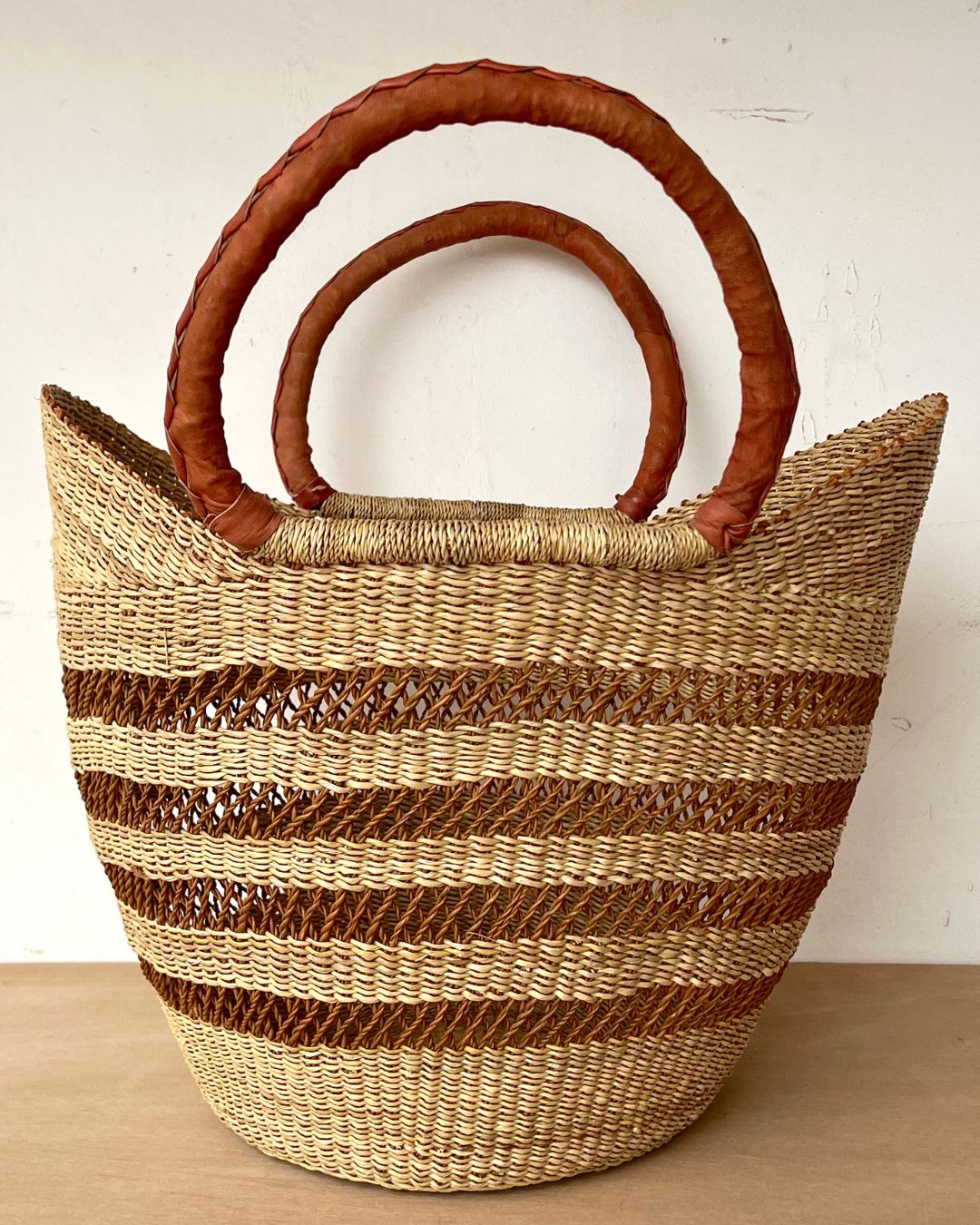 A woven straw tote bag with horizontal brown stripes and two thick, curved brown leather handles, set against a plain off-white wall and light wood surface.