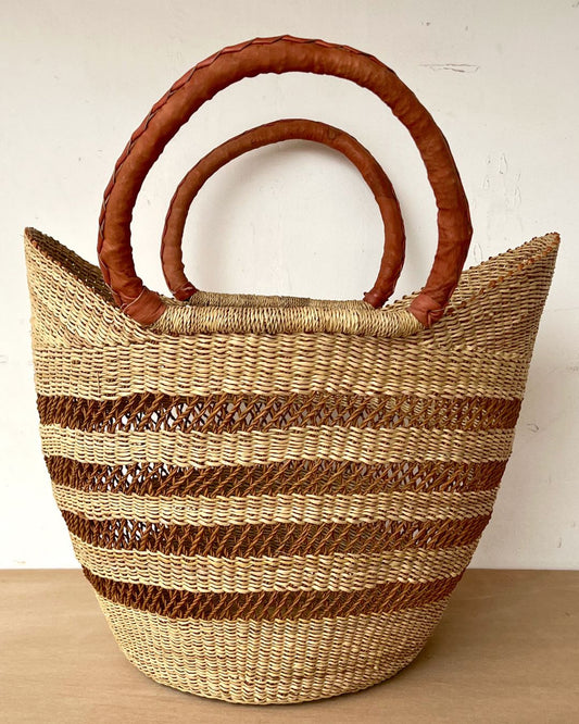 A woven straw tote bag with horizontal brown stripes and two thick, curved brown leather handles, set against a plain off-white wall and light wood surface.