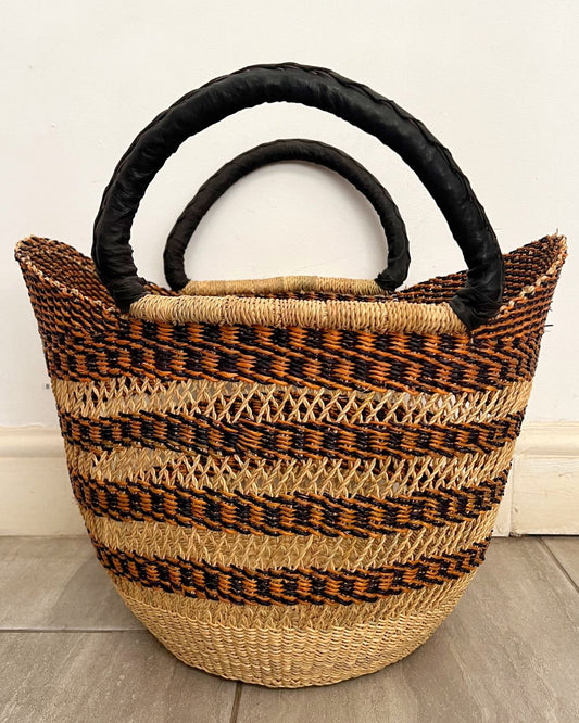 A woven straw tote bag with black handles, featuring horizontal stripes of natural beige, brown, and black raffia on a light gray floor against a white wall.