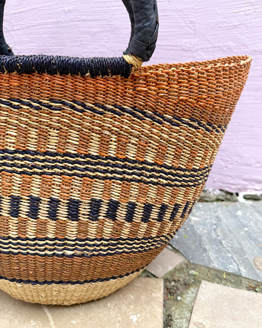 A woven straw tote bag with horizontal stripes in natural beige, brown, and black, featuring dark brown leather handles, set against a light purple wall and stone floor.