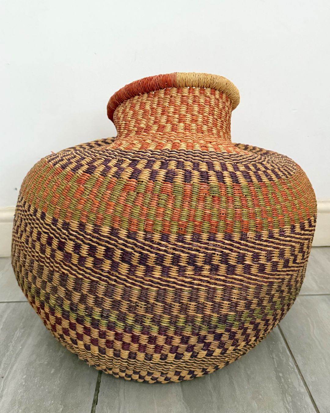 A large hand-woven basket vase with a bulbous body, narrow neck, and flared rim, featuring a geometric pattern in natural tan, brown, orange, and muted green on a light gray floor against a white wall.