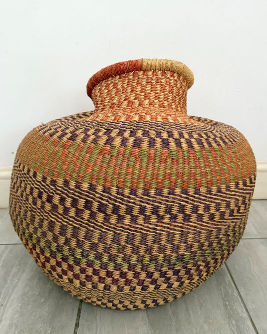 A large hand-woven basket vase with a bulbous body, narrow neck, and flared rim, featuring a geometric pattern in natural tan, brown, orange, and muted green on a light gray floor against a white wall.