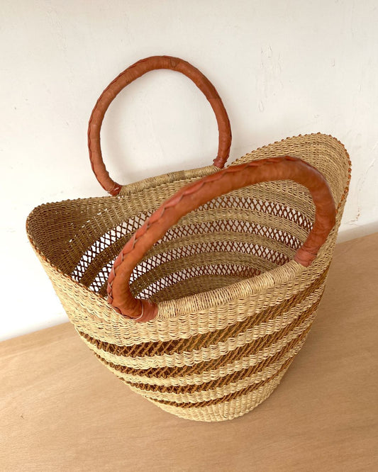 A woven straw tote bag with two curved brown leather handles, resting on a light wooden surface against a white wall.