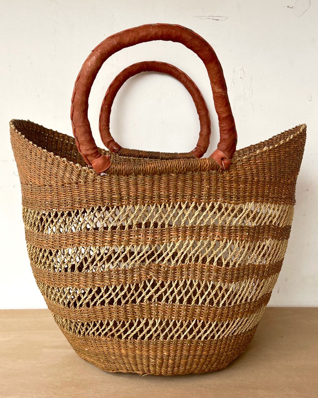 A woven straw tote bag with horizontal striped patterns in natural tan and light brown, featuring two thick, curved brown leather handles, set against a plain off-white wall and light wood surface.