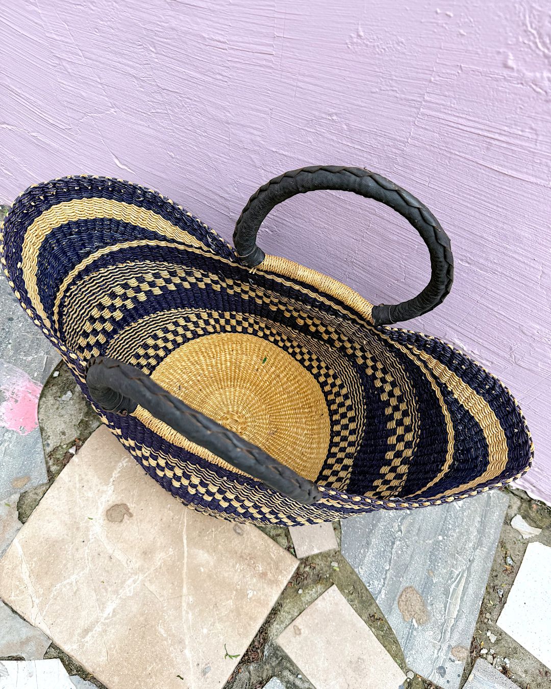 A woven straw basket with black handles, featuring a striped pattern of navy blue, black, and natural tan, resting on a stone surface against a light purple wall.