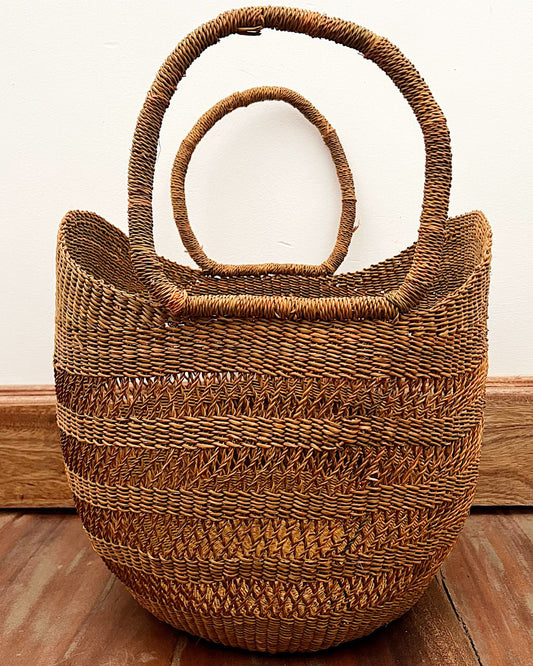 A large woven straw tote bag with two loop handles, shown on a wooden floor against a plain off-white wall.