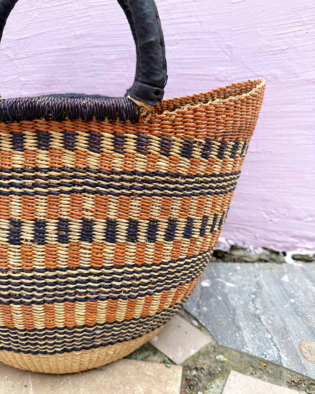 A woven basket bag with a curved black handle, featuring horizontal stripes of orange, beige, and black. It is photographed outdoors against a light purple wall and stone tiles.