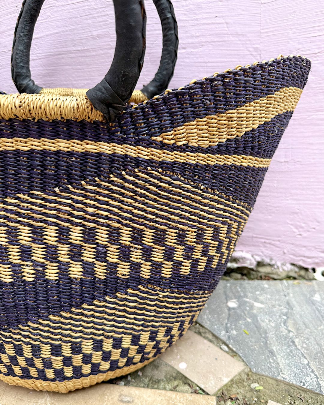 A close-up of a woven basket bag with dark navy and natural tan stripes, featuring dark brown leather handles, set against a light pink wall and stone floor.